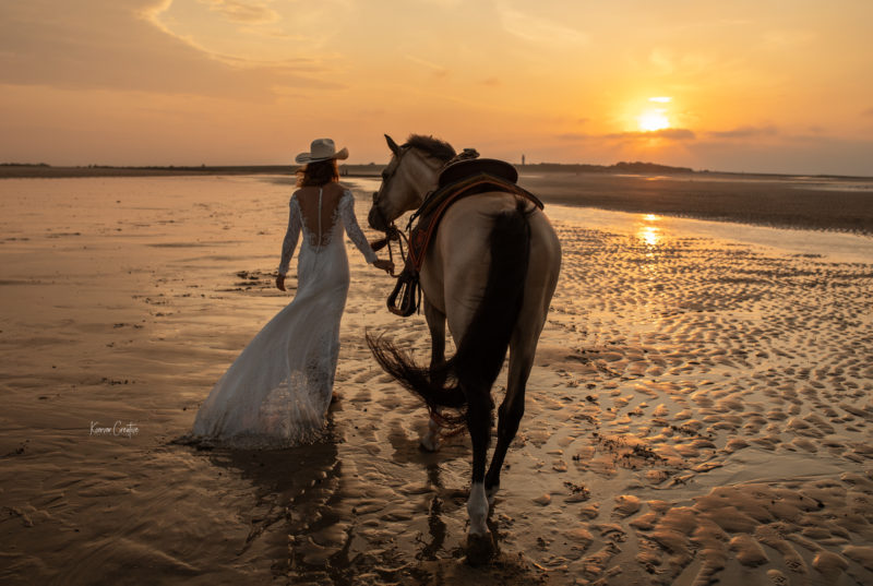 Pferd und Frau in Brautkleid mit Westernhut am Strand im Sonnenuntergang