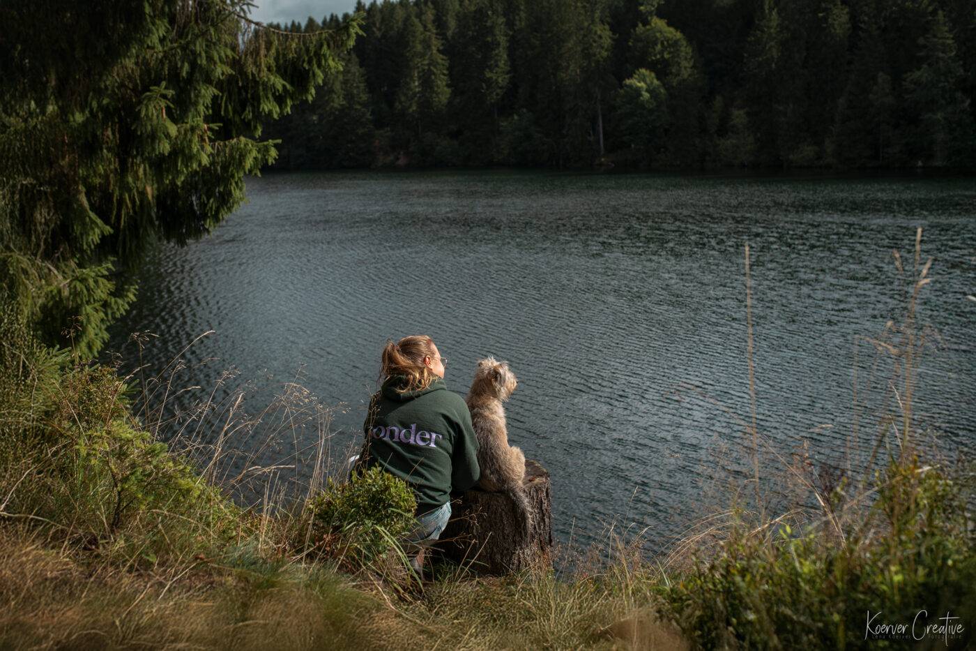 Eine Frau sitzt mit einem kleinen Hund an einem Badesee im Harz